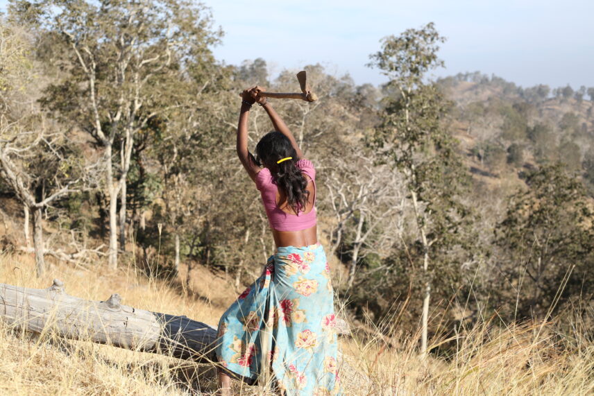 The photograph features a woman, with her back turned towards us. She is wearing a pink top and a light blue floral dress. She is chopping wood with an axe. In the background, we can see trees and hills.