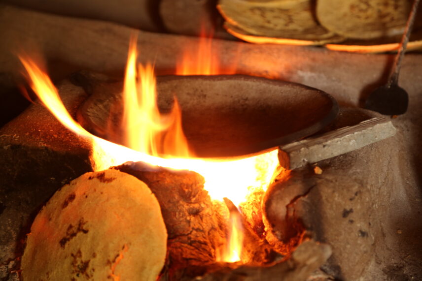 A close-up photograph of a mud stove. We can see the fire burning and baked naan in the background and in the front.