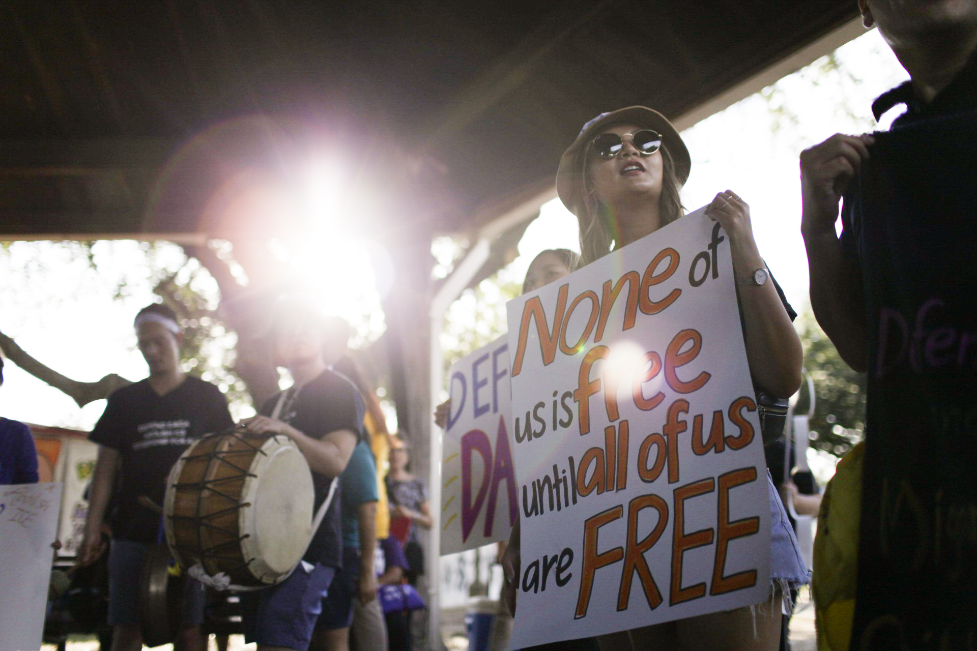 Protesters holding drums and signs, one sign reading "None of us is free until all of us are free."