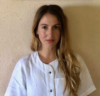 A photo of an individual standing against a tan wall and wearing a white shirt with long hair pushed over one shoulder.