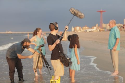 Two children and two adult actors stand in the edge of the ocean on a beach. A film crew with camera and sound boom film them.