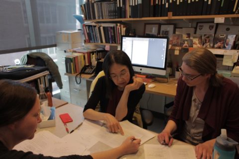 Three women sit at a table in an office with computer equipment and notebooks looking at documents together.