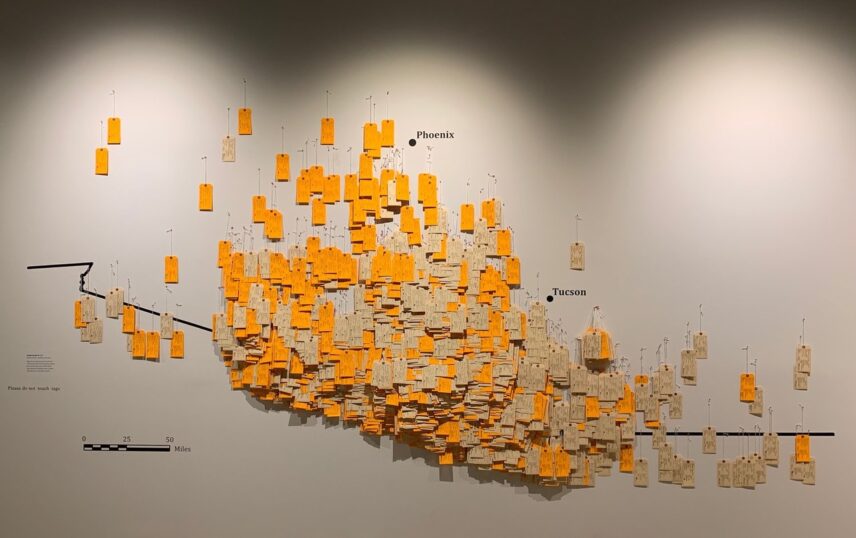 Yellow tags are attached to a white gallery wall. The tags are arranged to create a map of the Arizona-Mexico border.