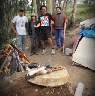 A photo of four people standing in the background with trees behind them. The foreground includes a small fire, caribou hand drum, and part of what appears to be tent.