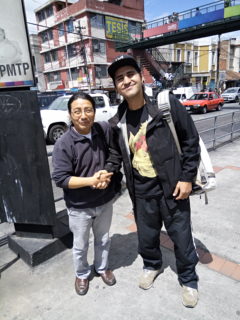 A photo of two people smiling at the camera and shaking hands on a city sidewalk.
