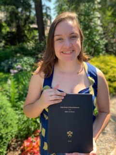 A photo of an individual with long brown hair smiling at the camera and holding a book in front of greenery.