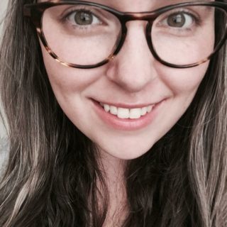 A close-up photo of an individual smiling at the camera and wearing tortoise-shell patterned glasses, brown hair visible.