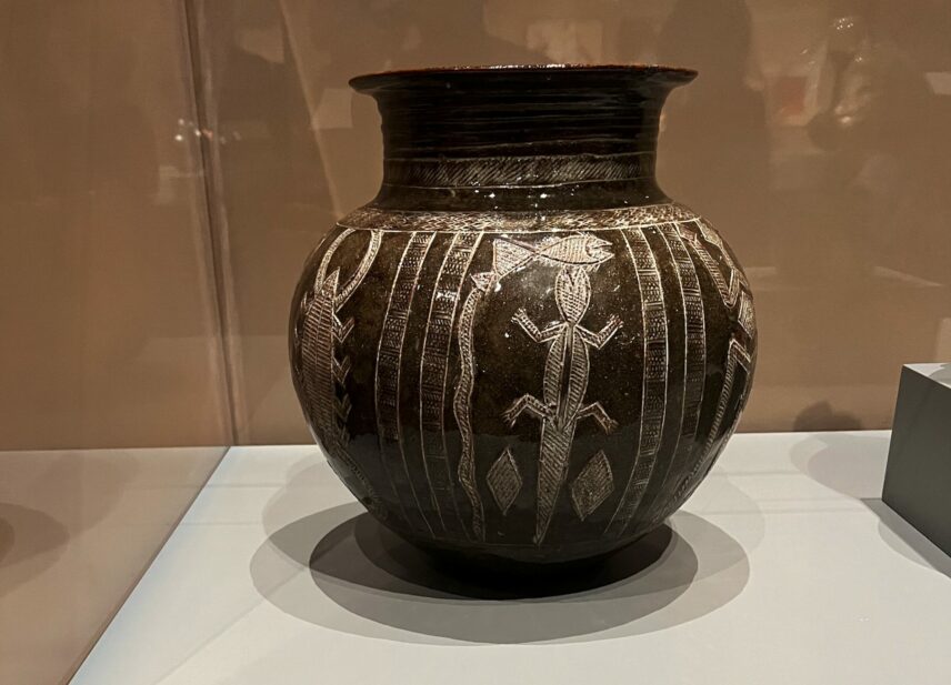 A globular glazed stoneware vessel with incised decoration filled with white pigment on display behind plexiglass.