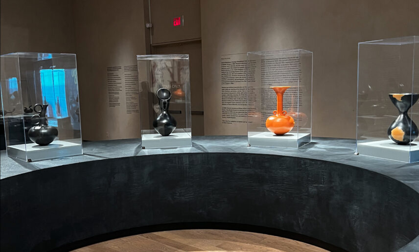 Four ceramic vessels in plexiglass cases sit atop a large, black, semi-circular ceramic platform.