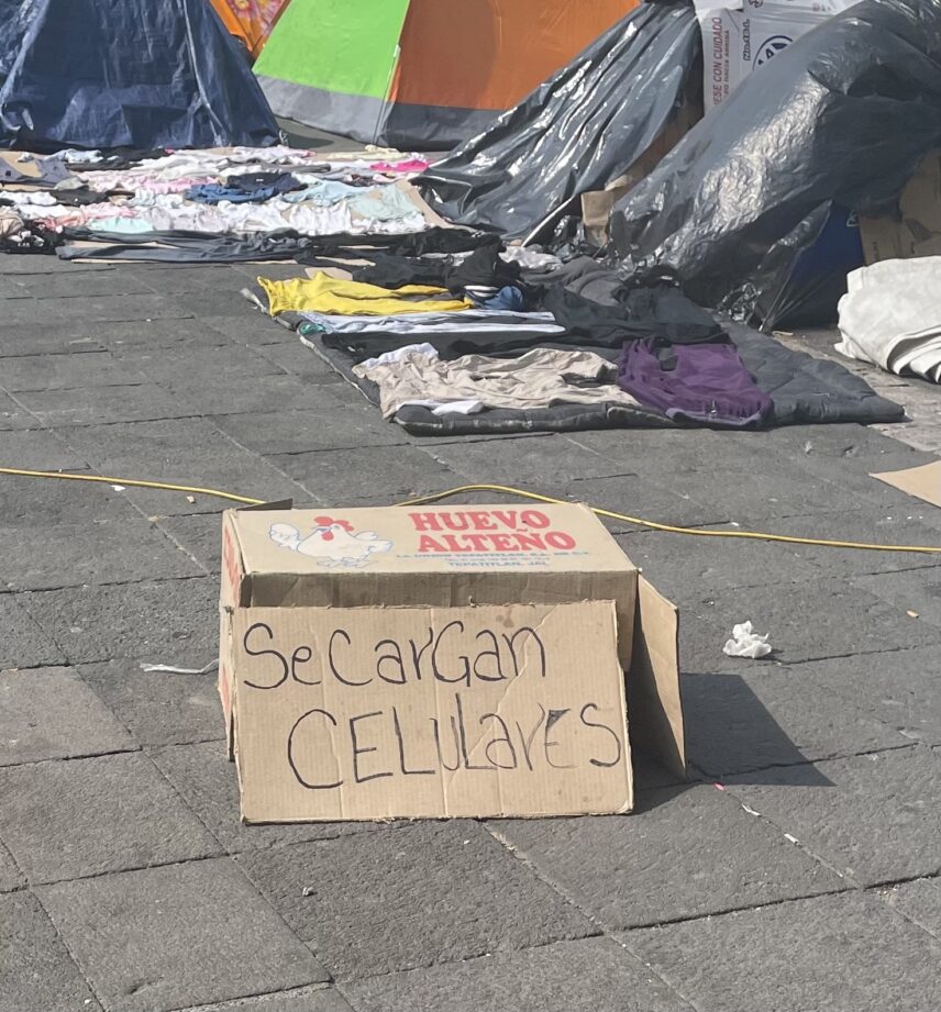 A handwritten sign on a repurposed box of chicken eggs reads: "Cell phones are charged here". The sign is in a migrant camp in Mexico City. Tents, and clothing lying on top of a tarp to dry, are in the background.